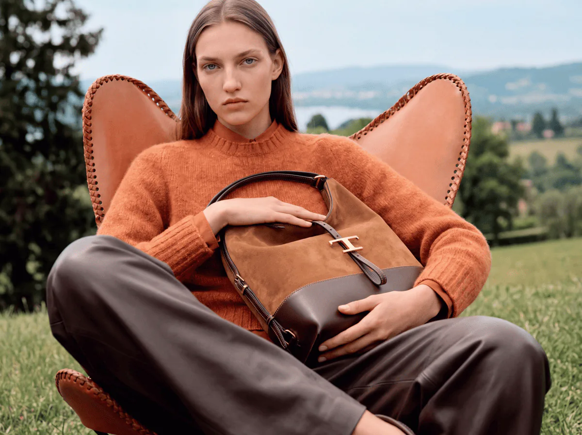 Le même mannequin virtuel en campagne : pull orange, pantalon en cuir, un sac en daim à la main, dans un fauteuil en cuir sur une colline