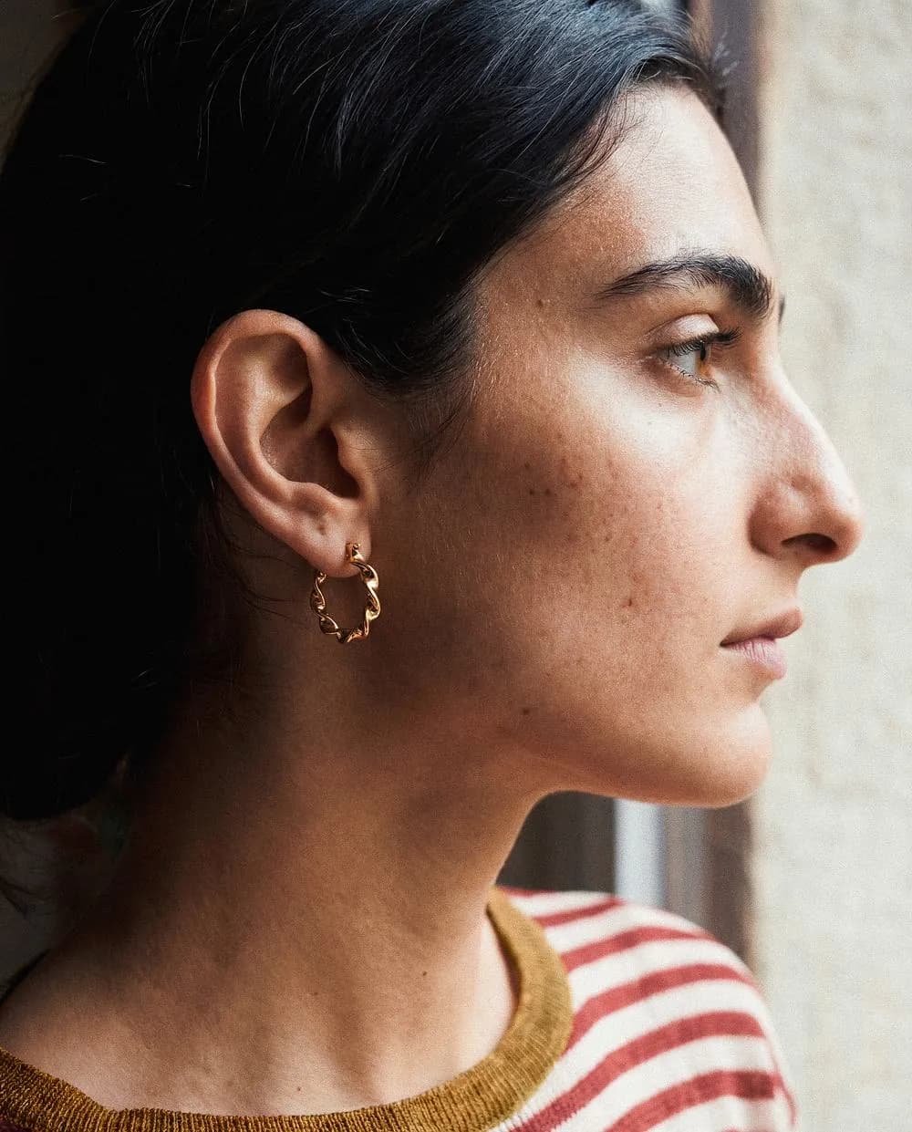 Profile of a model by a window, wearing a gold hoop earring and a striped sweater