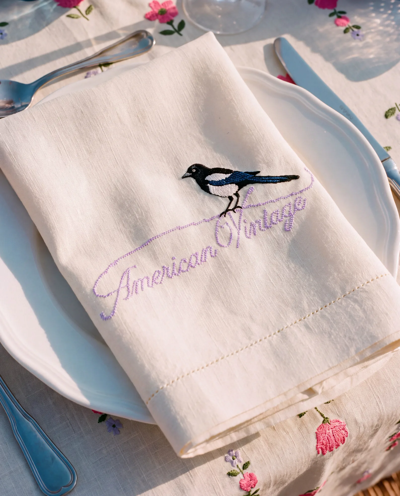 Linen napkin embroidered with a magpie and the American Vintage logo on a floral tablecloth