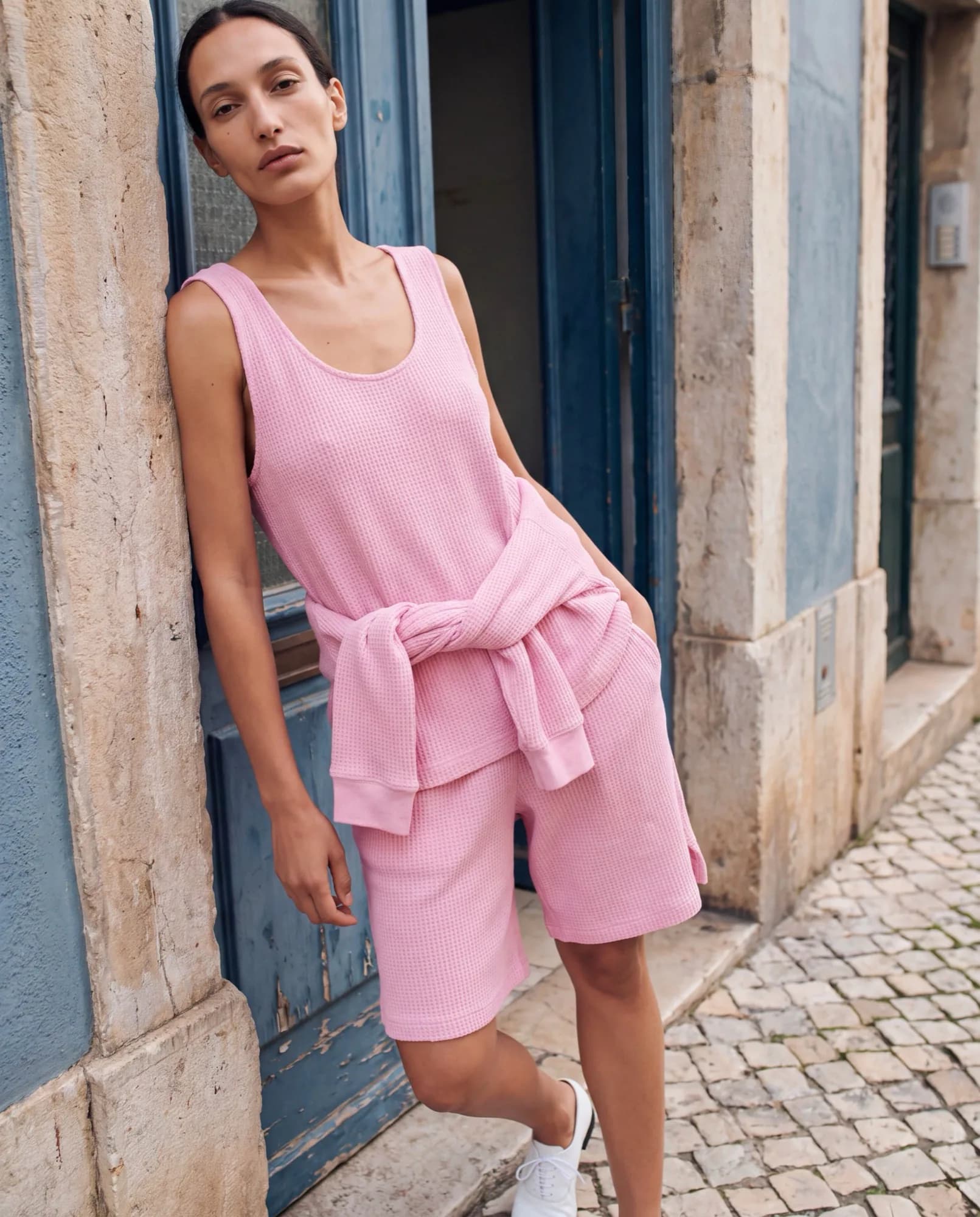 Model in a pink tank top and shorts with a sweater tied at the waist, leaning in a stone doorway