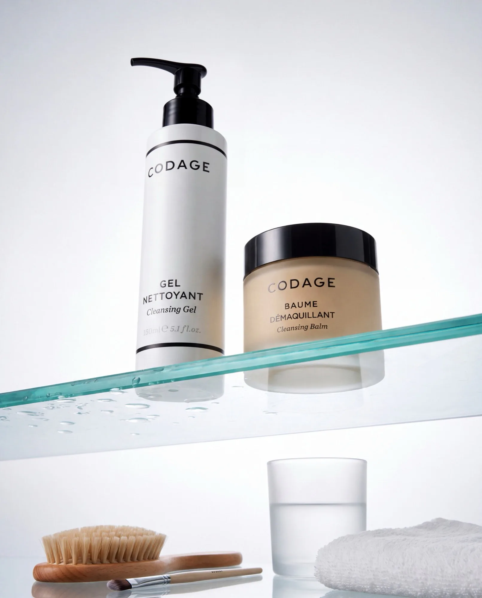 Codage cleansing gel and cleansing balm on a glass shelf above a brush and a glass of water