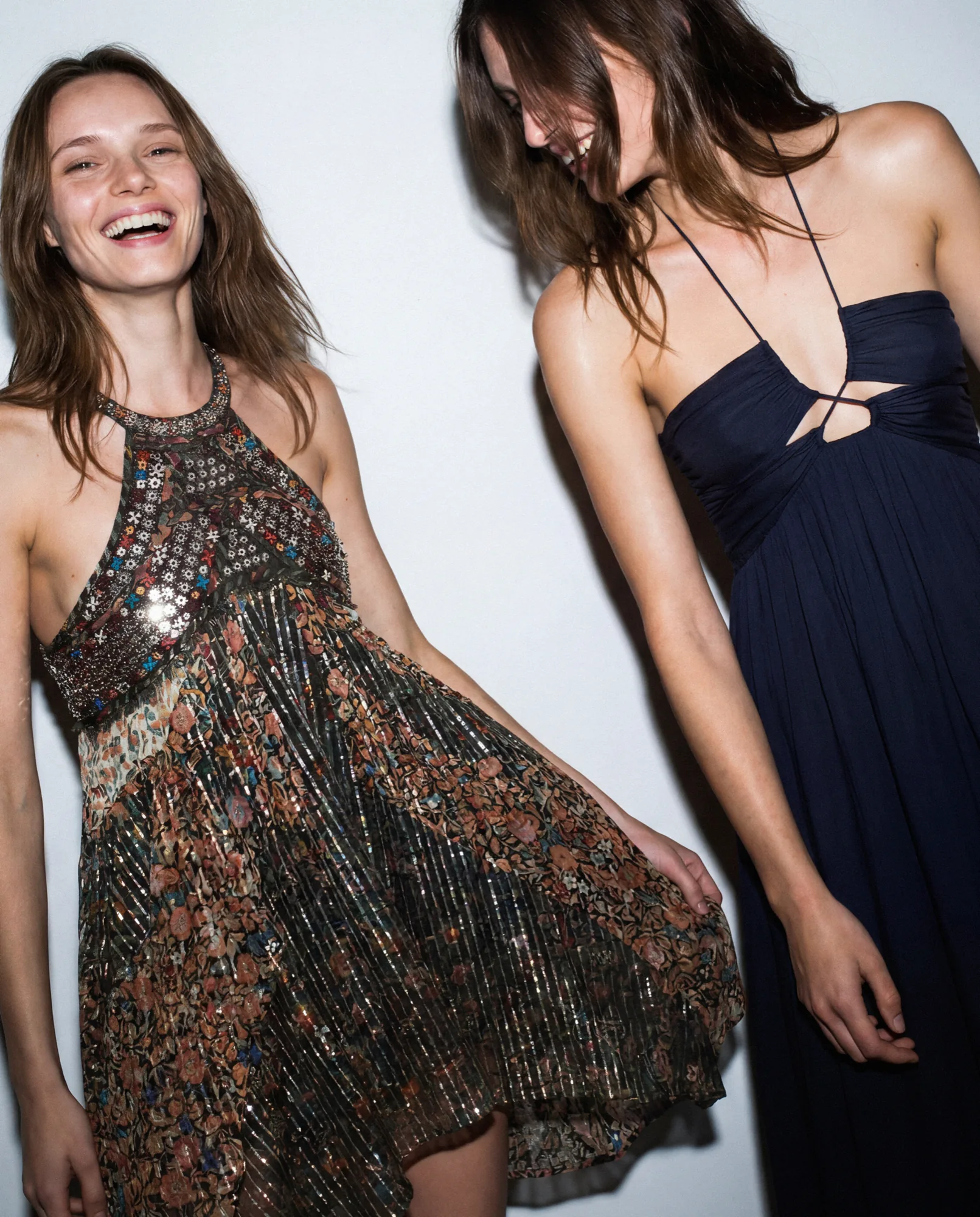 Deux mannequins qui rient, l’une en robe imprimée à sequins, l’autre en robe dos nu marine