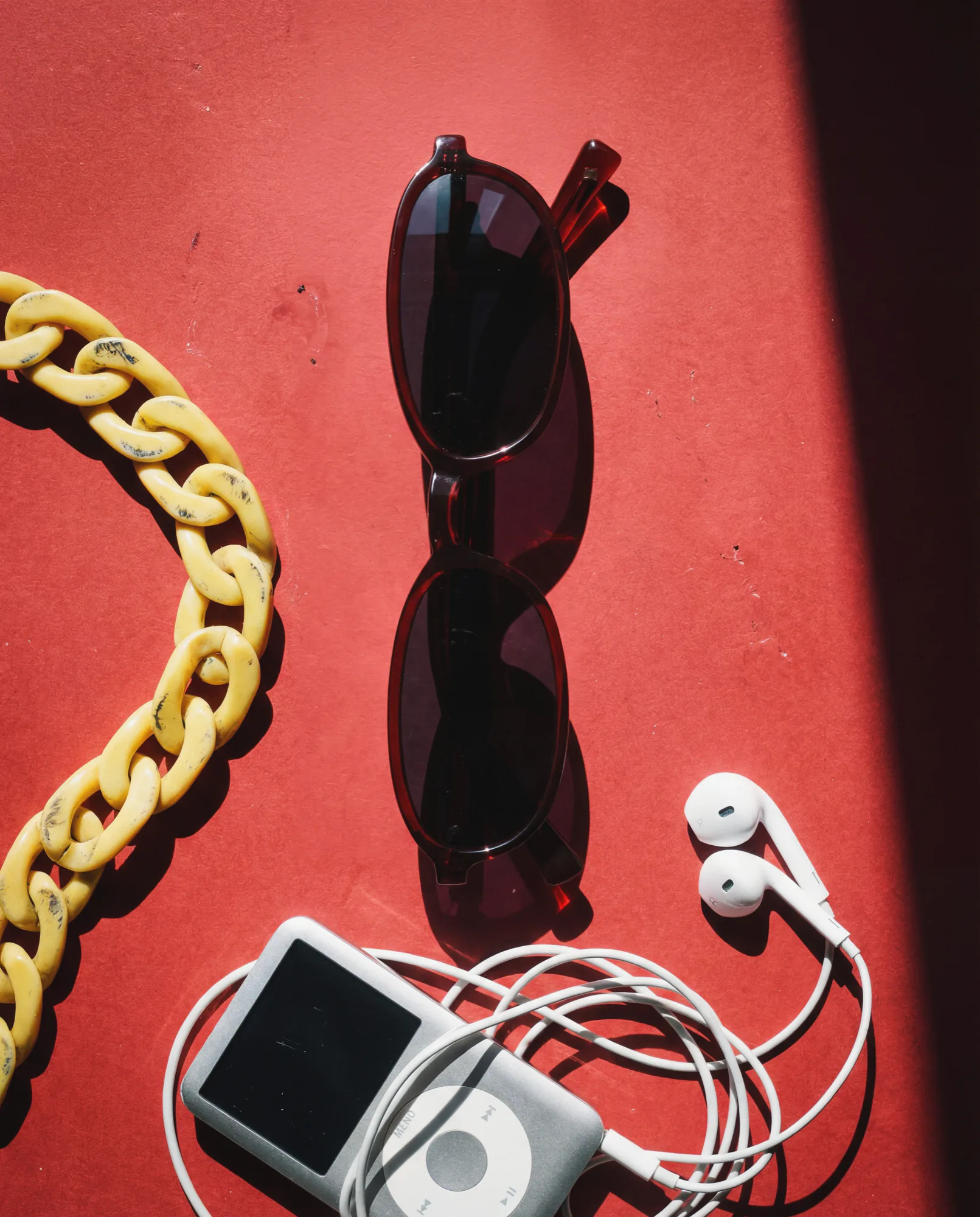 Lunettes de soleil foncées sur une surface rouge, à côté d’un gros bracelet chaîne et d’un baladeur MP3 avec écouteurs