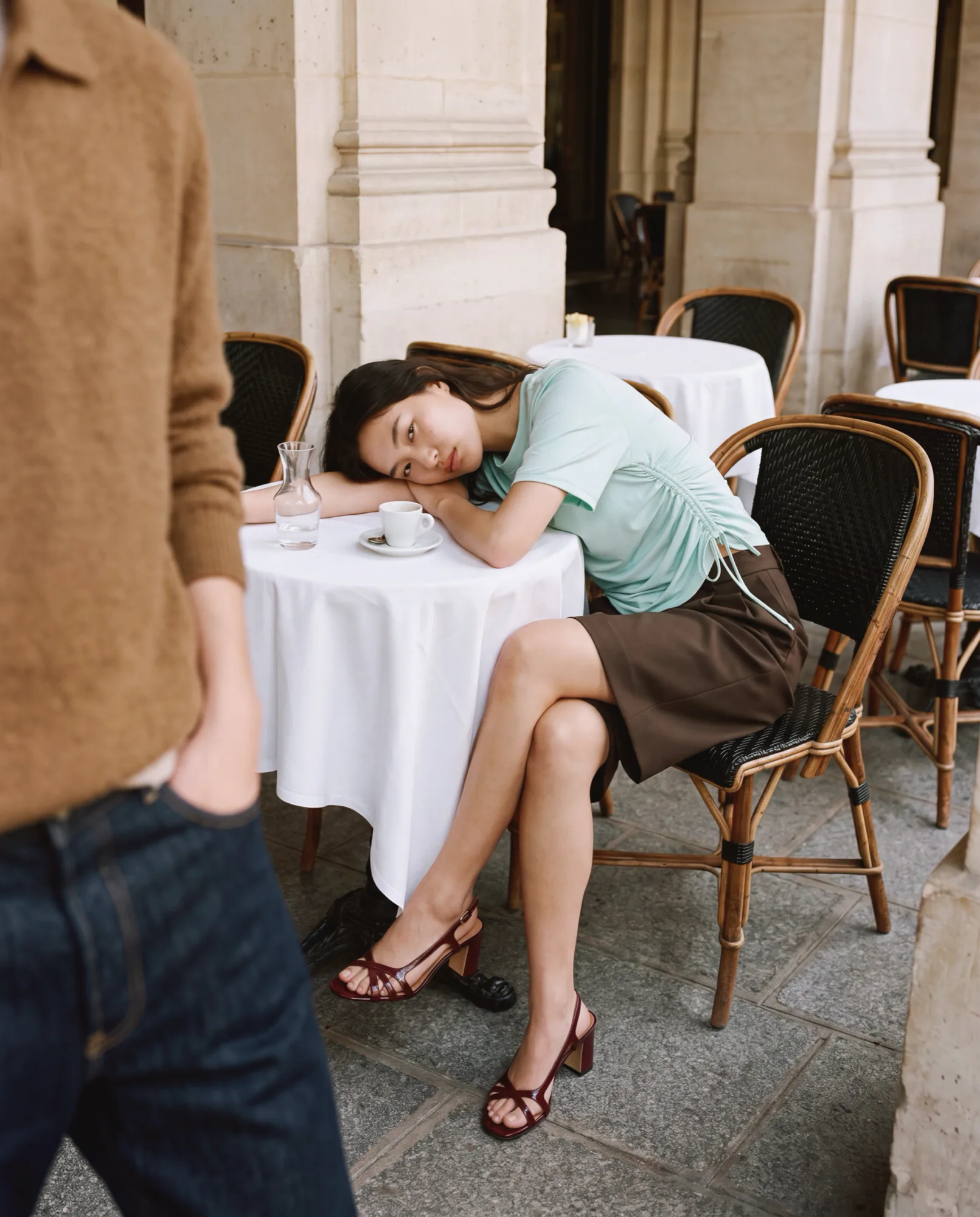 Model resting her head on a café table on a Paris terrace, wearing strappy heeled sandals
