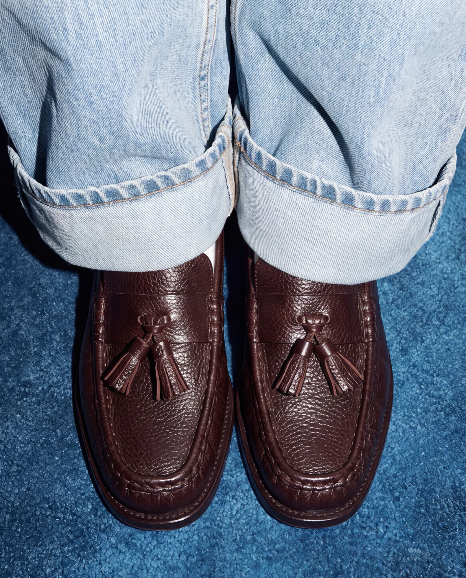 Burgundy tassel loafers under rolled-up jeans on a blue background