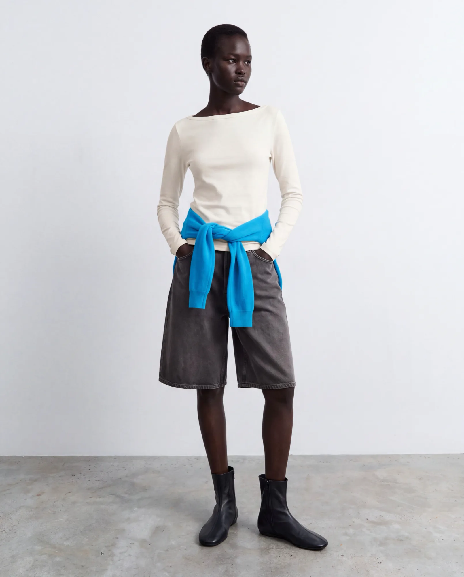 Model in a cream top, grey shorts and a blue sweater tied at the waist, with black ankle boots
