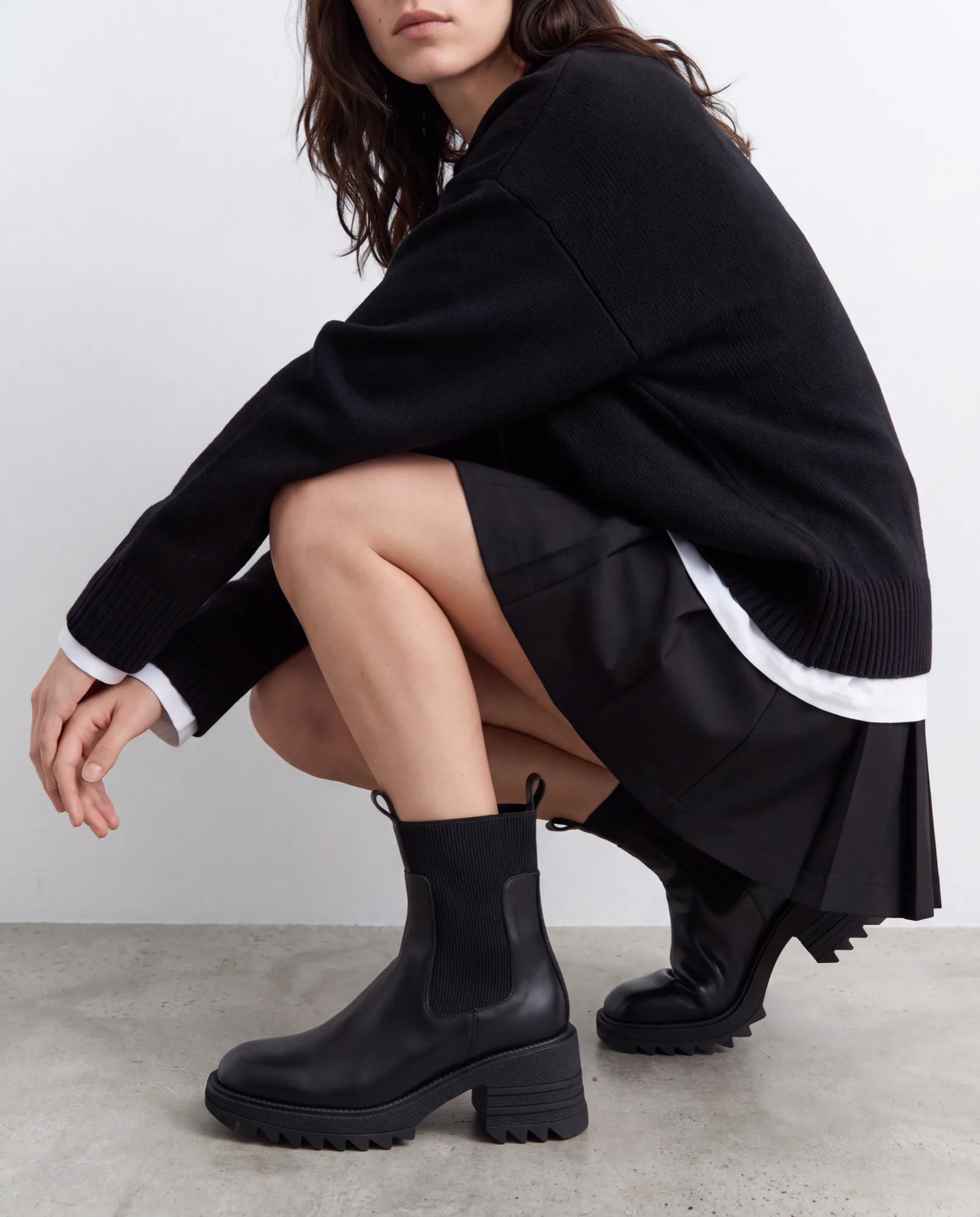 Mannequin accroupie en pull noir et jupe plissée, avec des boots noires épaisses