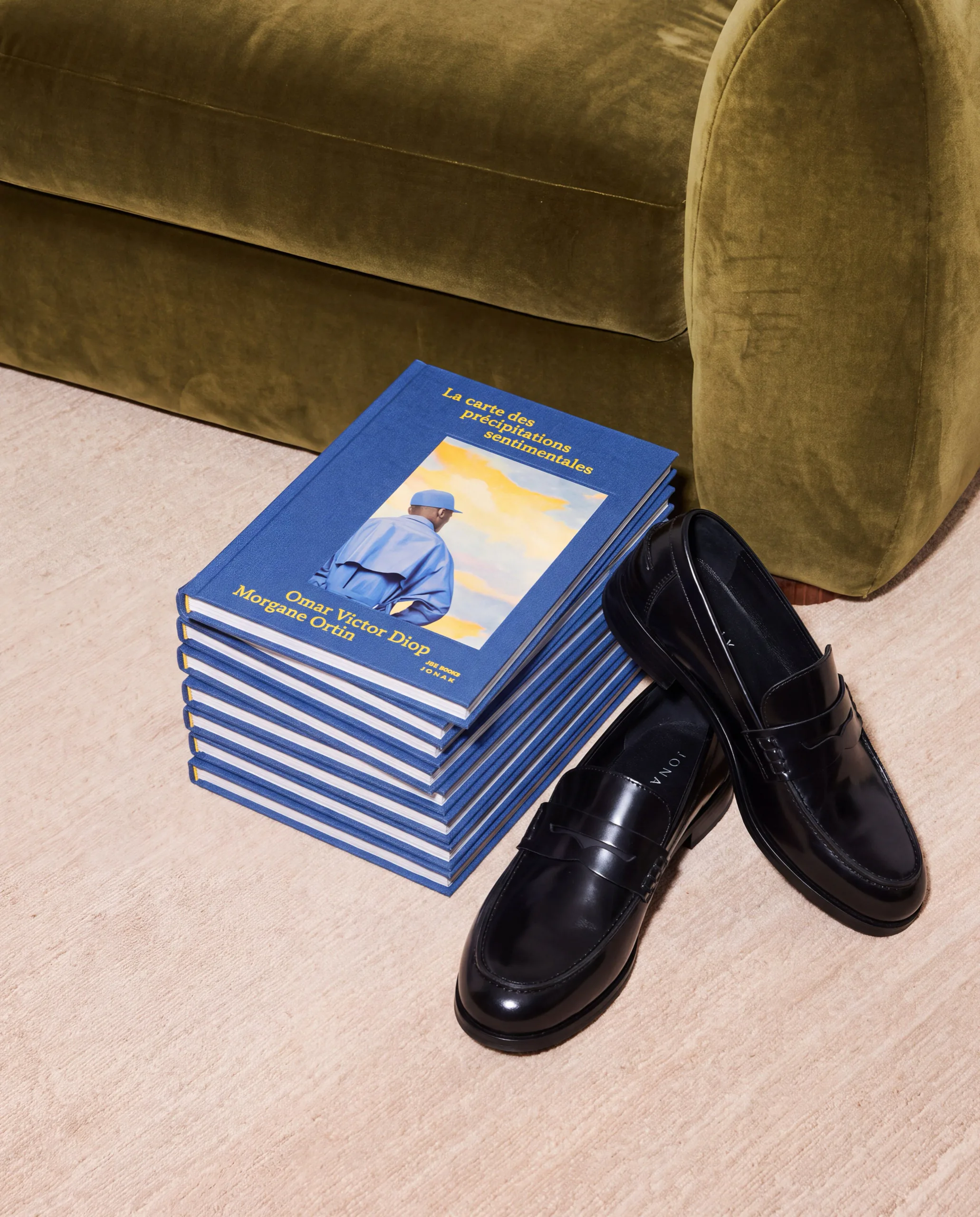 Black loafers next to a stack of blue books by an olive velvet sofa