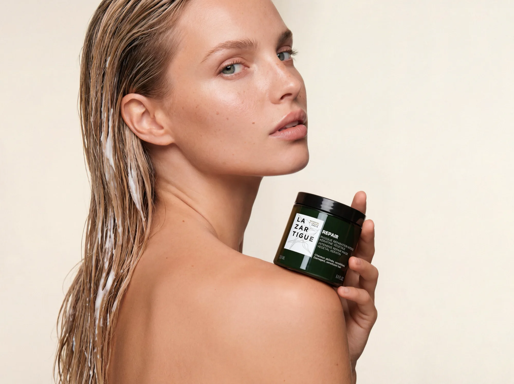 Model with wet slicked-back hair holding a jar of Lazartigue repair mask