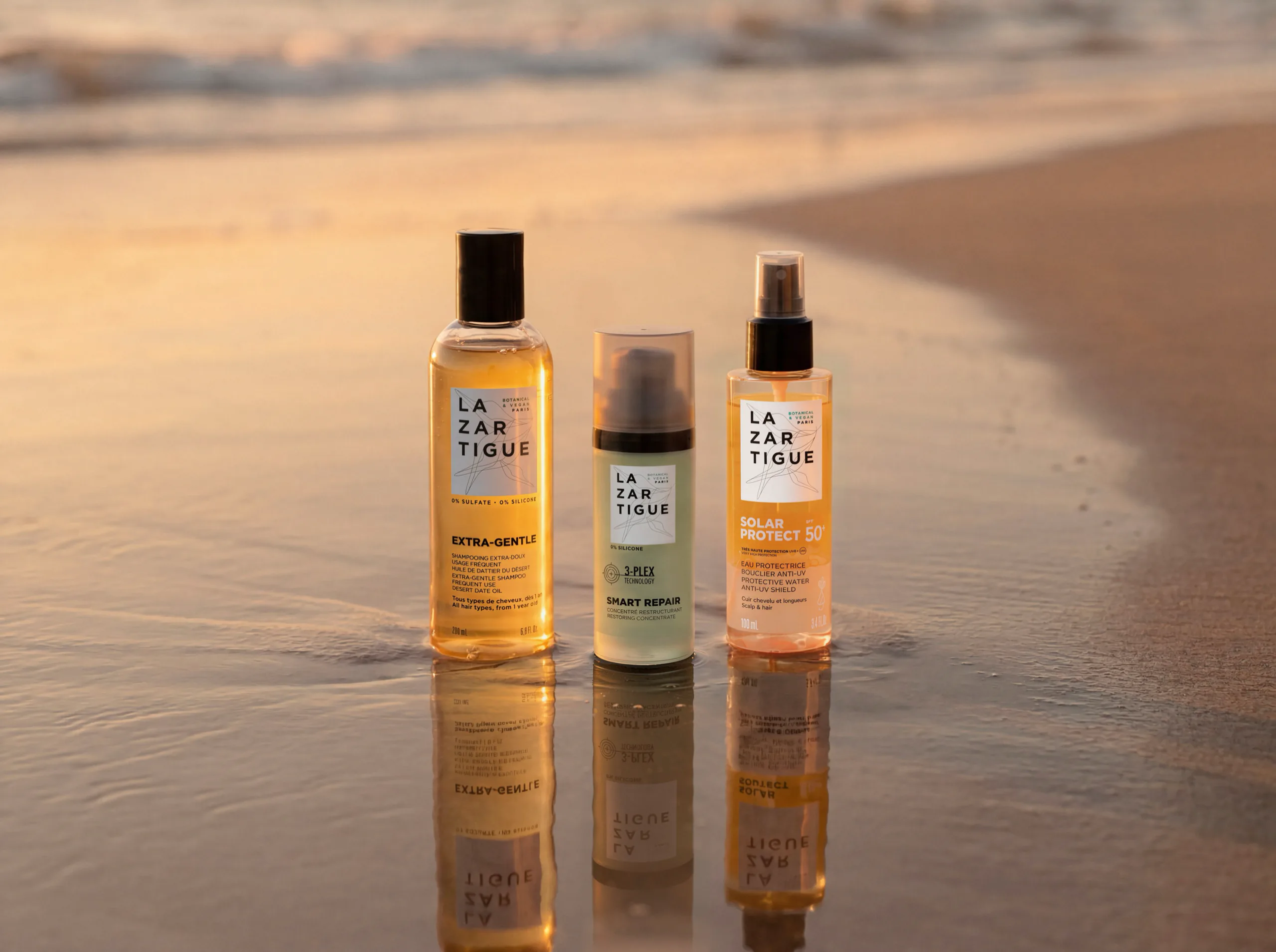 Three Lazartigue haircare products standing on wet sand at the water's edge at sunset