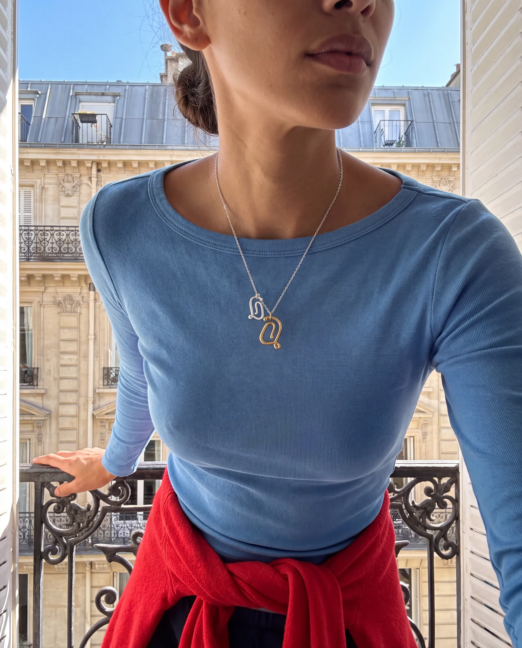 Femme en haut bleu portant un collier à pendentif monogramme sur un balcon parisien