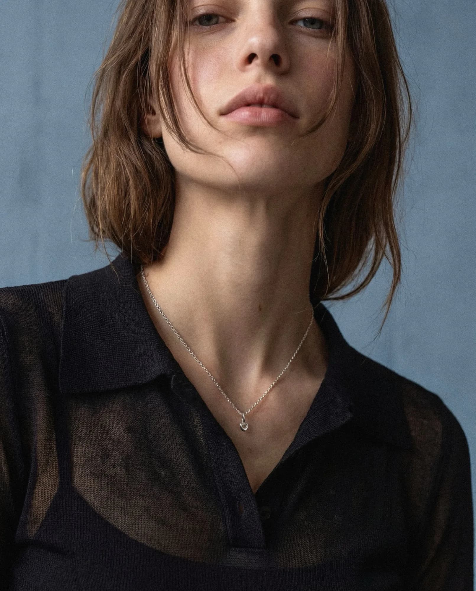 Model in a sheer black shirt wearing a fine silver necklace, against a blue wall