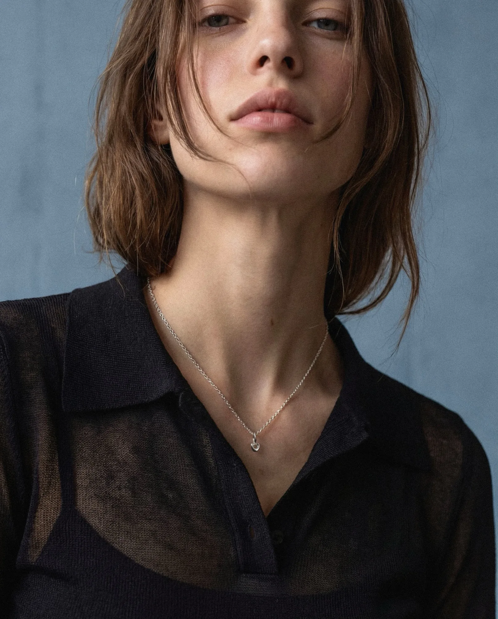 Model in a sheer black shirt wearing a fine silver necklace, against a blue wall