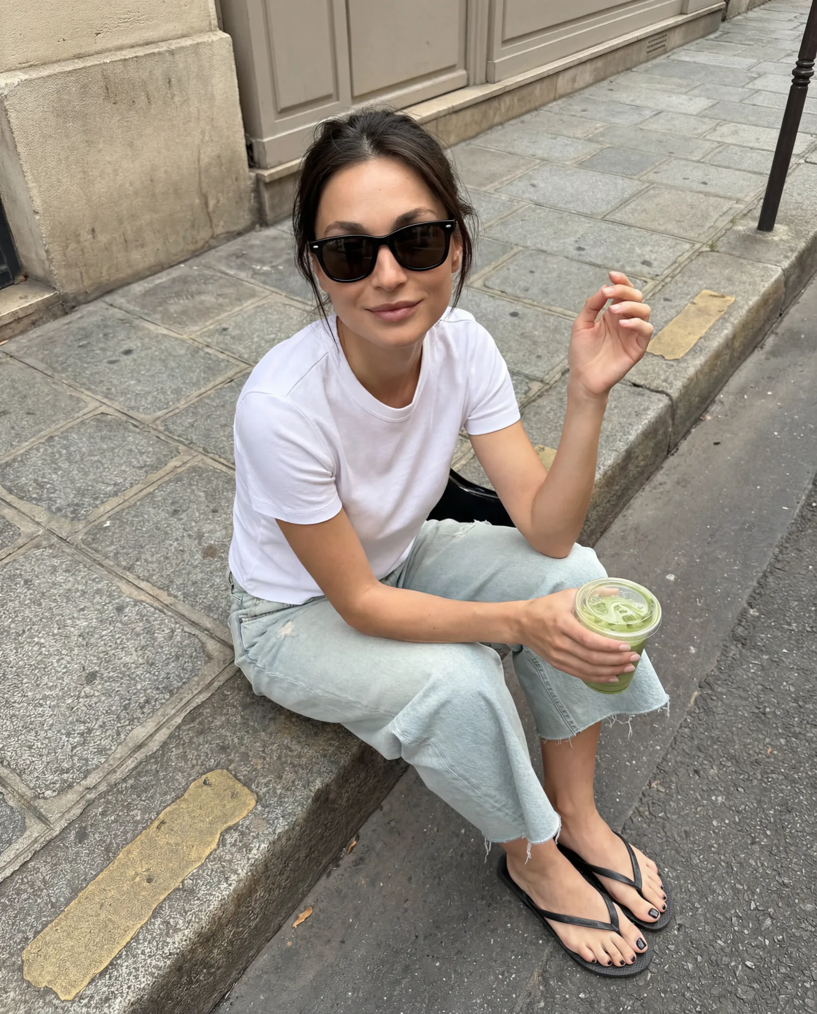 Woman in sunglasses sitting on a Paris sidewalk with a drink, UGC style