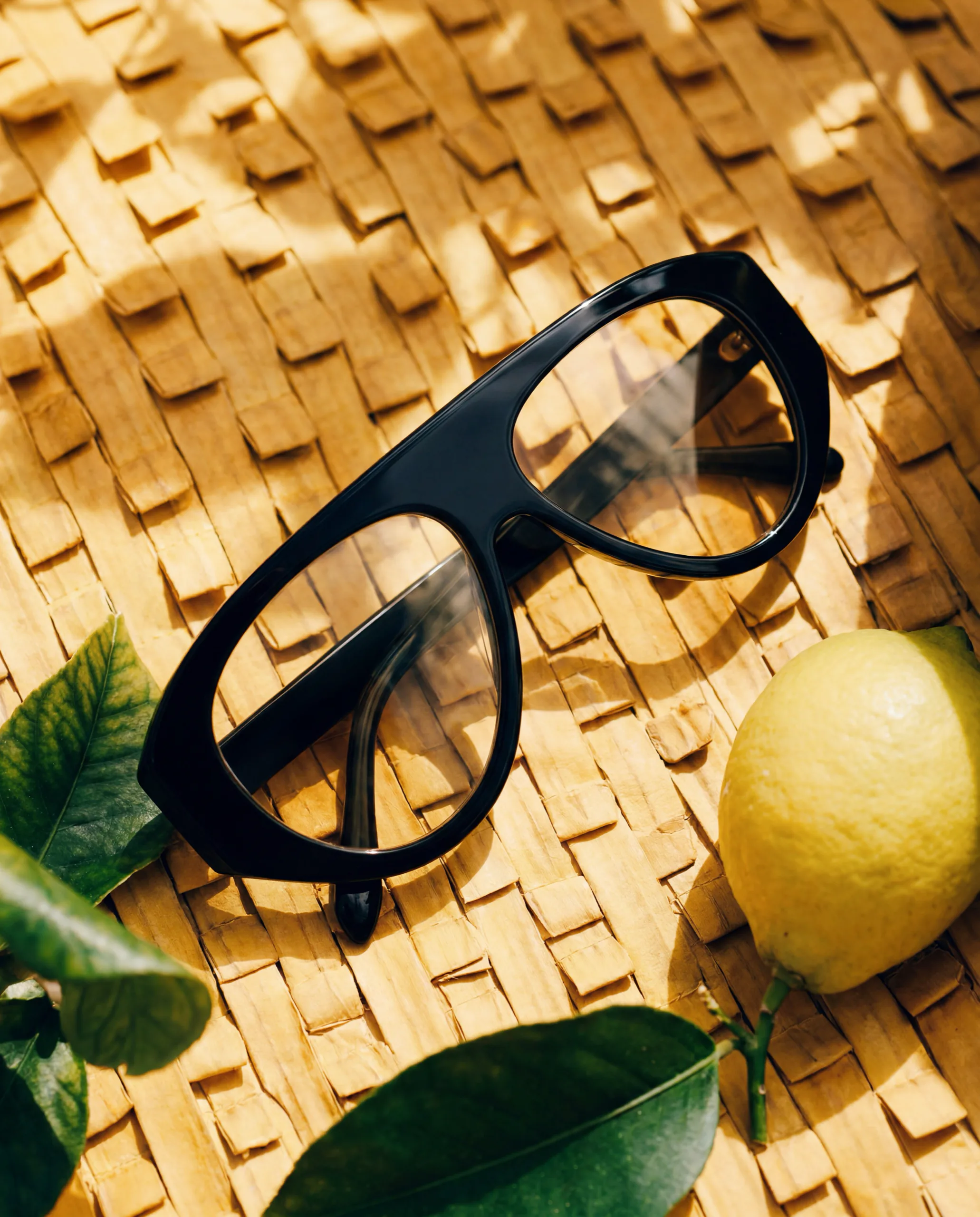 Black acetate glasses on a woven straw surface next to a lemon and leaves
