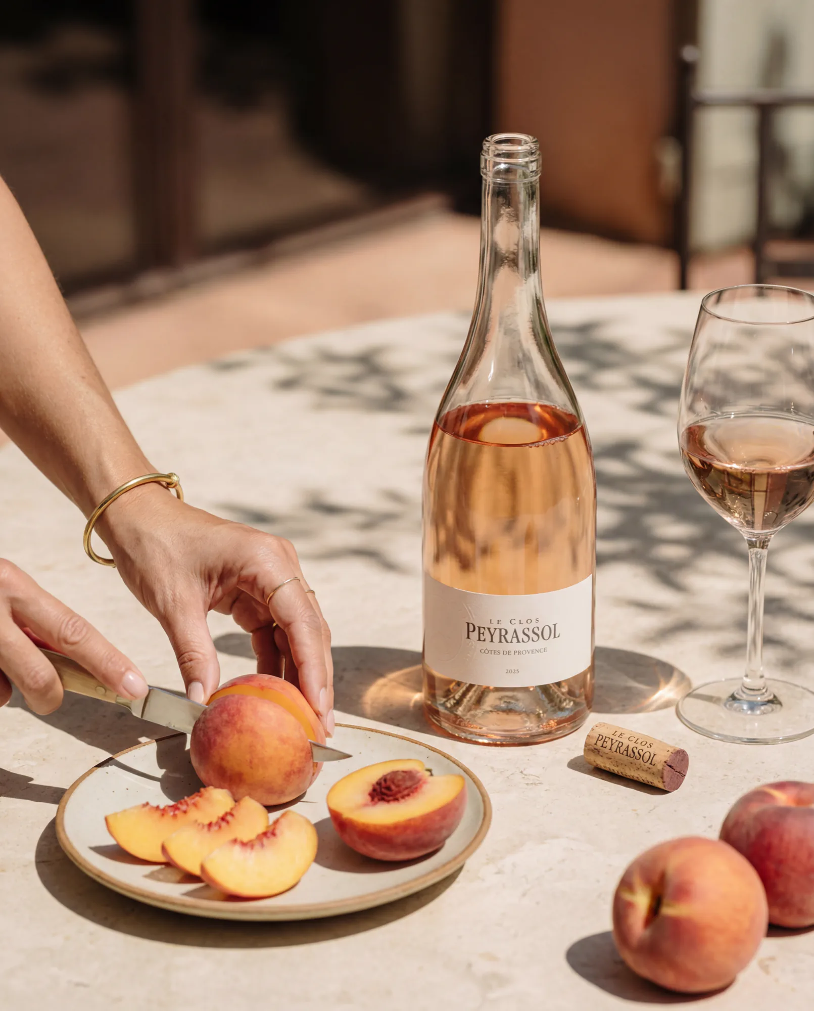 Mains coupant une pêche à côté d’une bouteille de rosé Peyrassol et d’un verre rempli