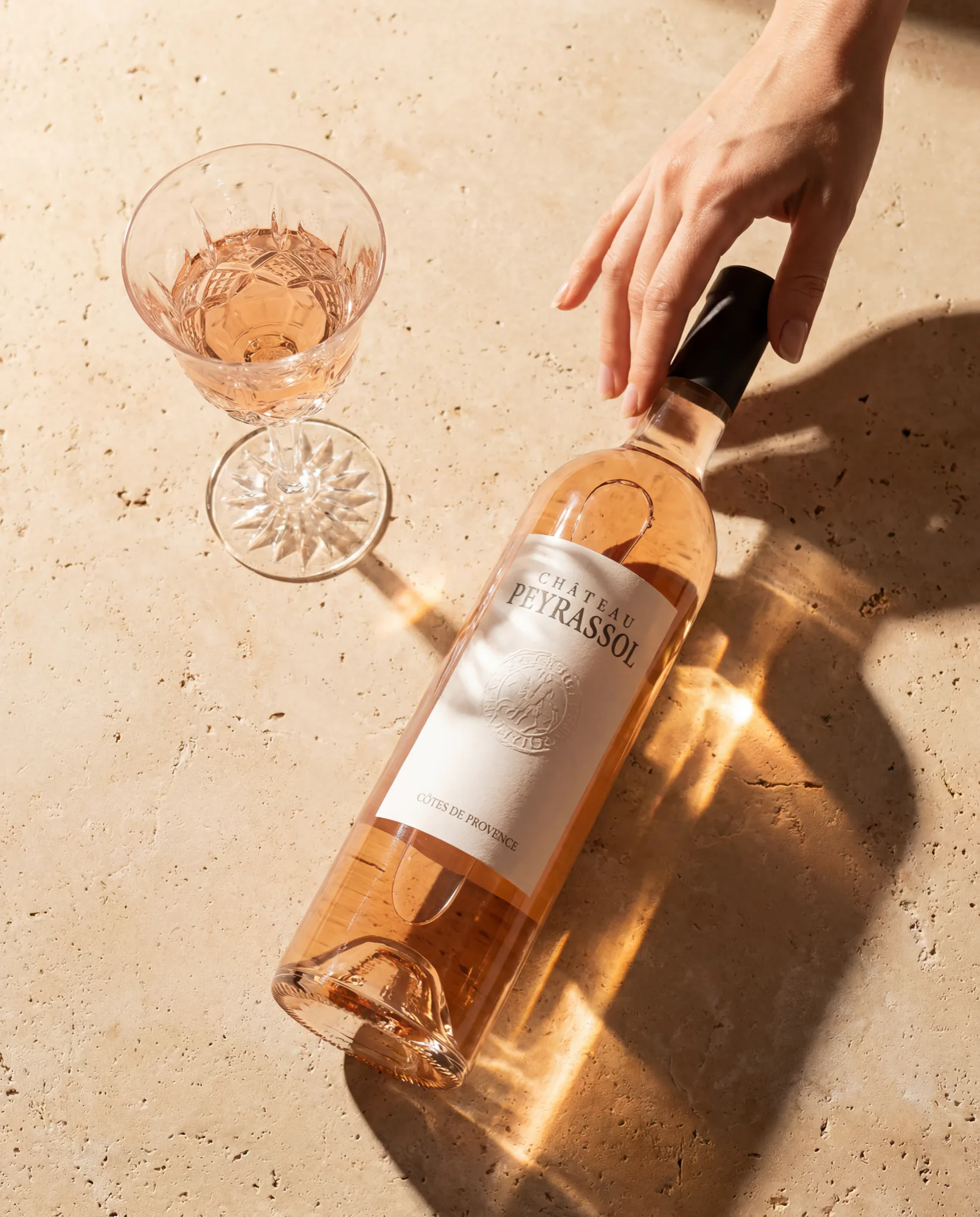 A hand laying a bottle of Peyrassol rosé on warm stone beside a cut-crystal glass