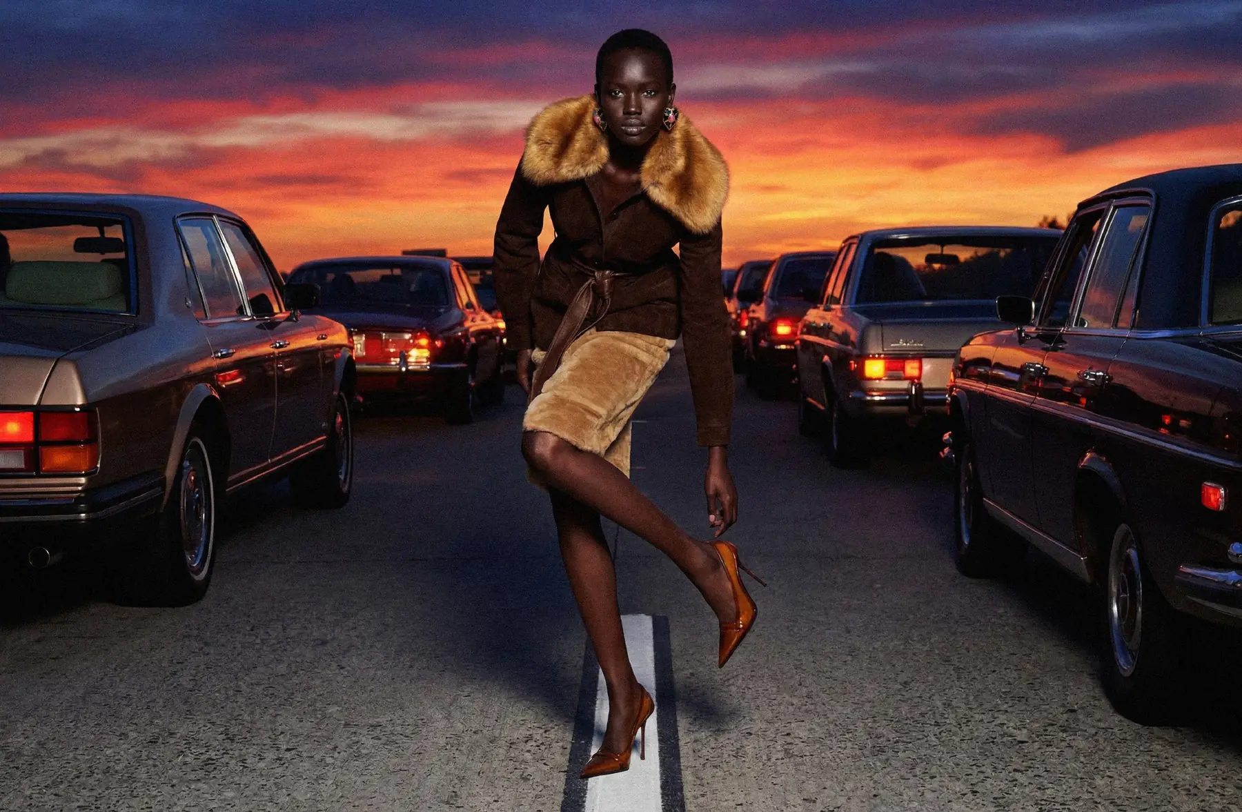 Model in a fur-collared coat on a road at sunset, between rows of vintage cars